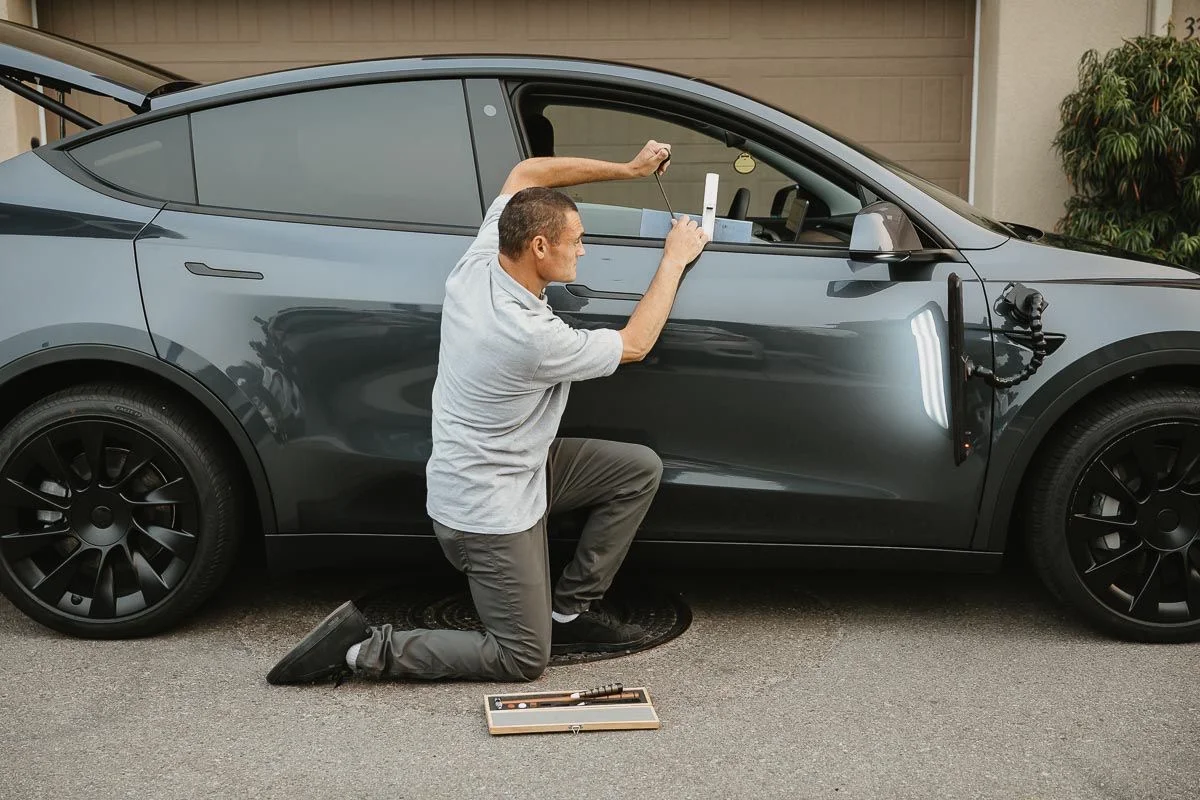 Technician kneeling beside a grey Tesla Model Y in a driveway, repairing a door dent