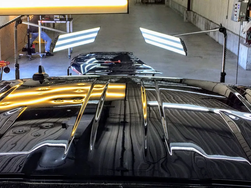 Hail dents showing in the reflection of a light board across a black roof, with the hood under repair behind