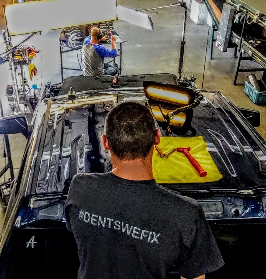 Technician working hail dents out of a dark car roof under a PDR light, with a second technician repairing the hood behind him