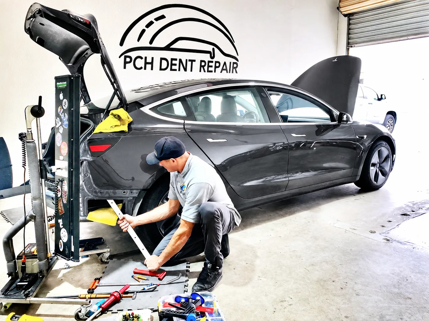 Technician repairing the rear quarter of a dark grey Tesla Model 3 with a PDR light board and tools